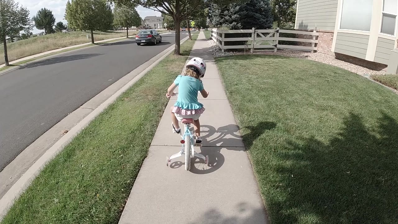At the start of the Colorado clip, a young girl in a white helmet rides away from camera on a sidewalk beside grass, a road, and suburban houses.
