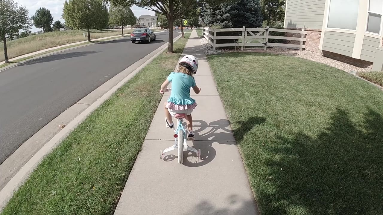 The girl continues riding away in Colorado, centered on the concrete path with the road on her left and a trimmed lawn on her right.