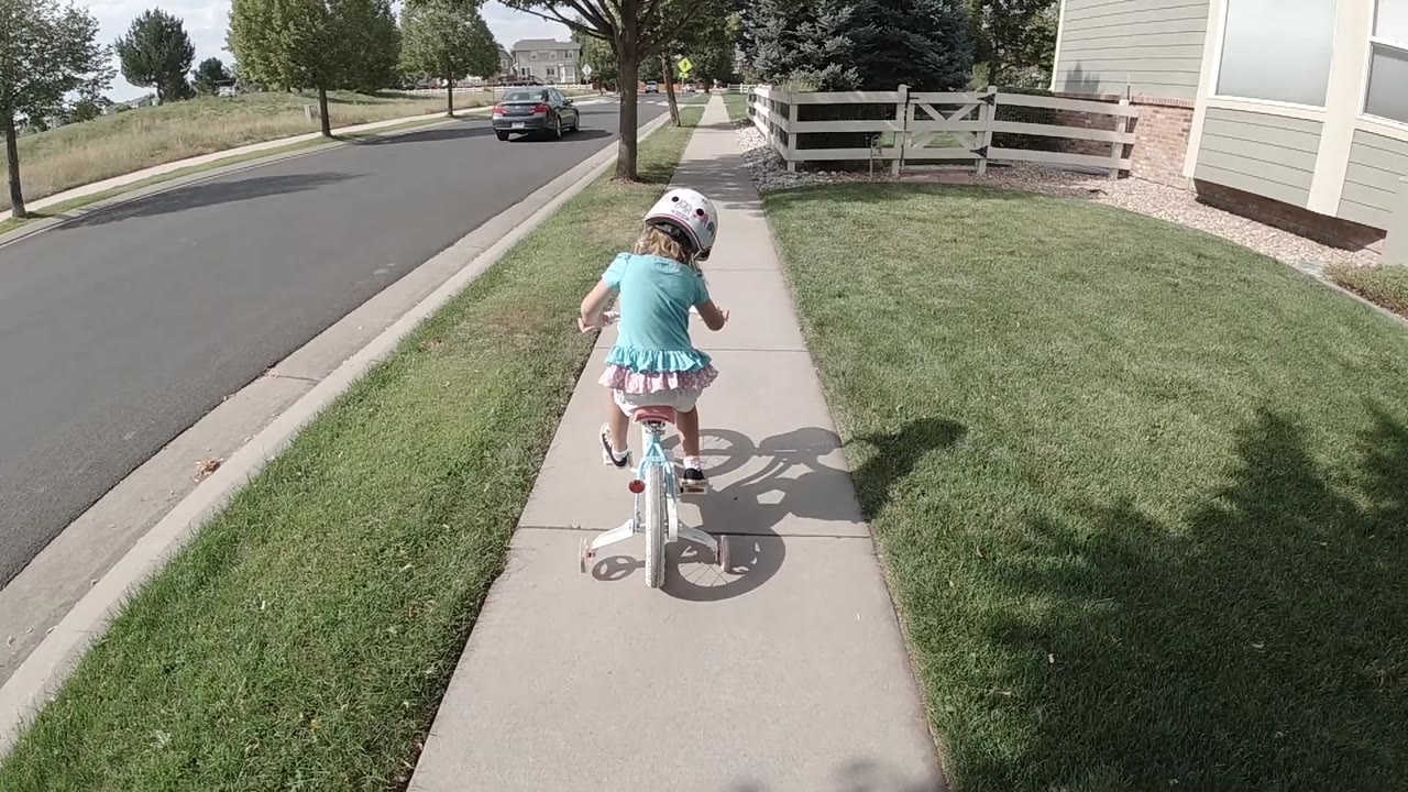 Girl pedals a small bike past grass and a house.