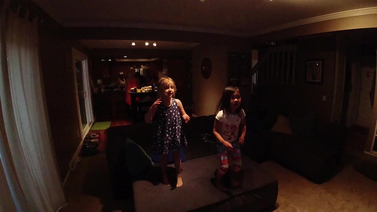 Two children stand near a dark sofa in a warm, dim living room at the start of the clip.