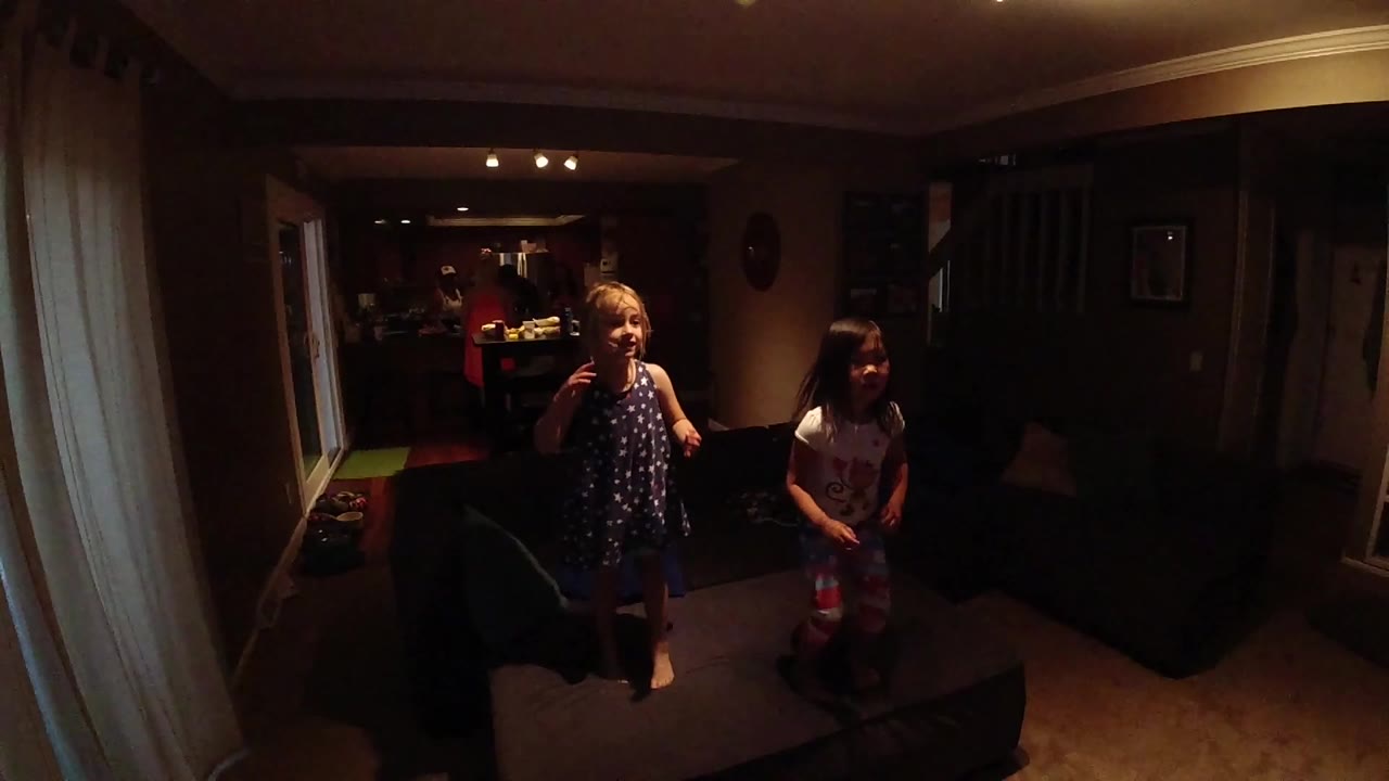 The two children face the camera from the center of the room, with the dark sofa and warm ceiling lights behind them.