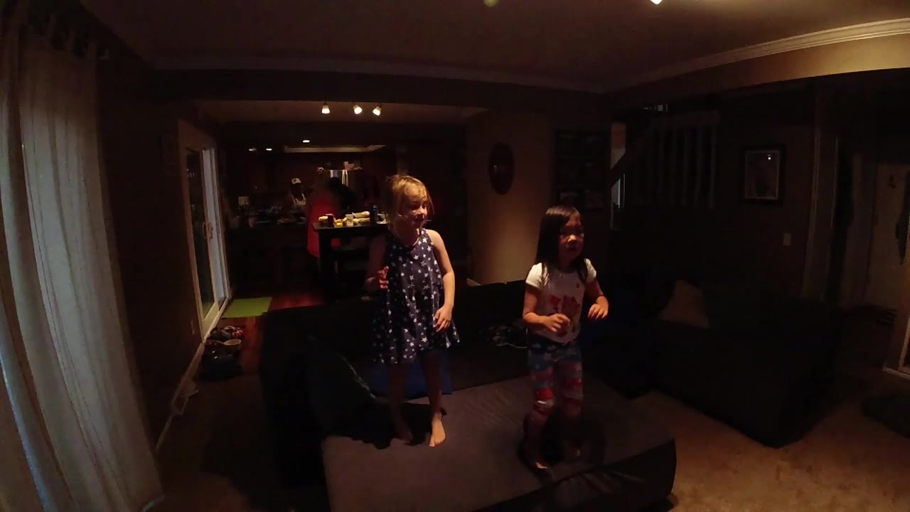 The children stand separated across the carpet, with the blond child at left and the child in a light shirt at right near the dark couch.