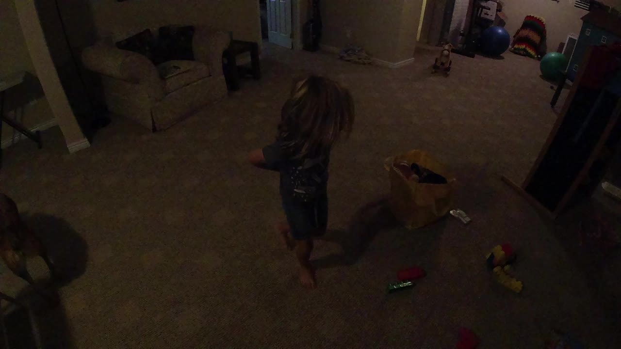 A young girl stands near scattered toys and a yellow bag in a dim carpeted living room at the start of the clip.