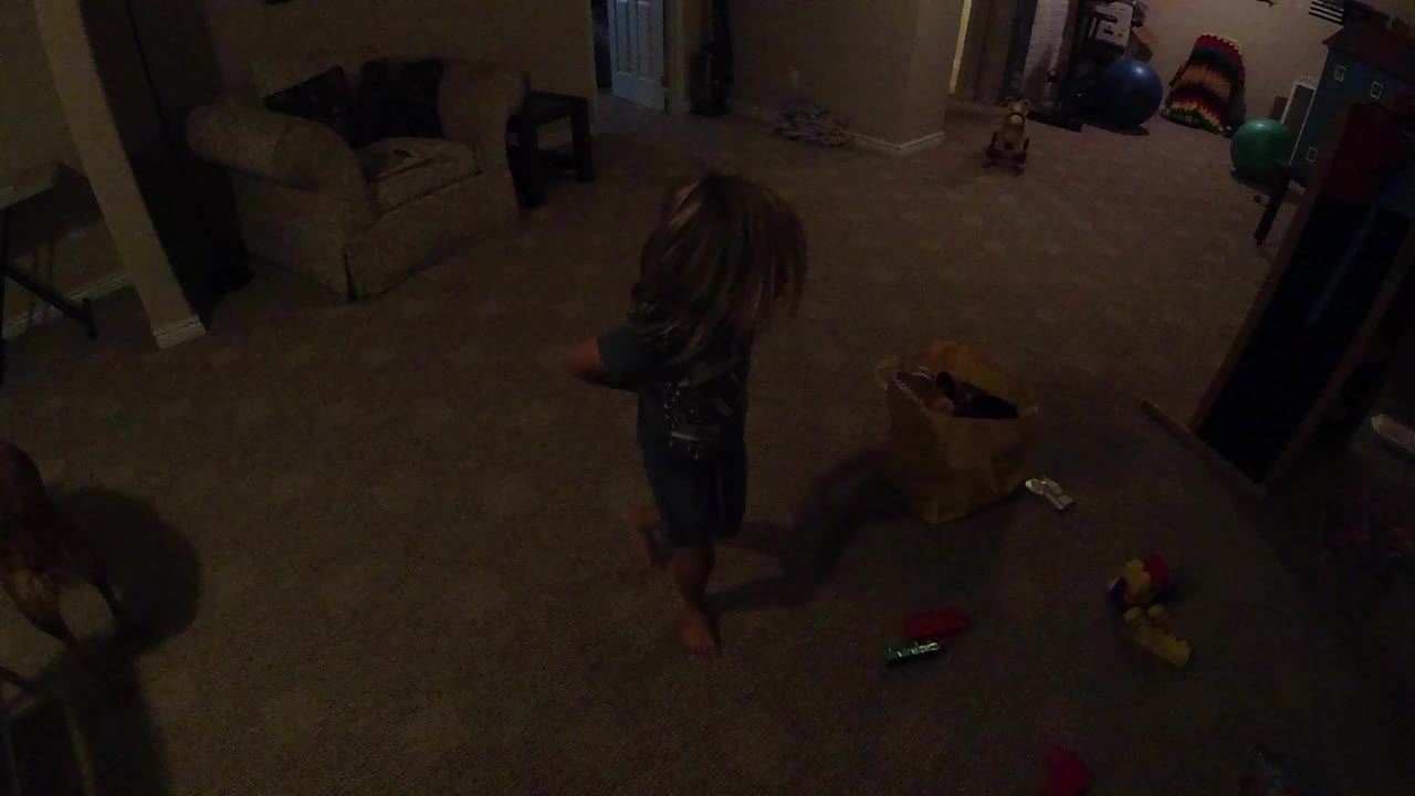 The girl remains near the yellow bag as toys lie across the carpet and furniture lines the darker room edges.