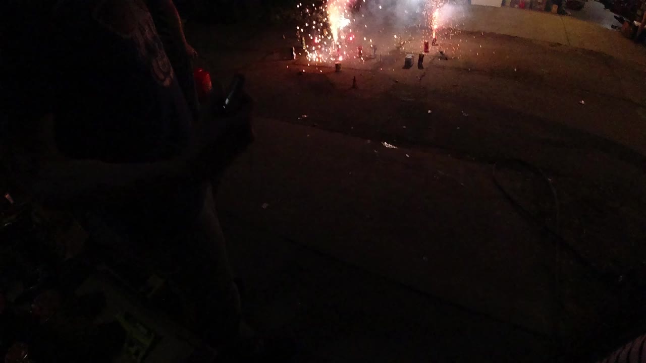 A red and white firework burns on a dark residential street in Denver collection footage, with shadowed people at the frame edges and an open garage in the background.