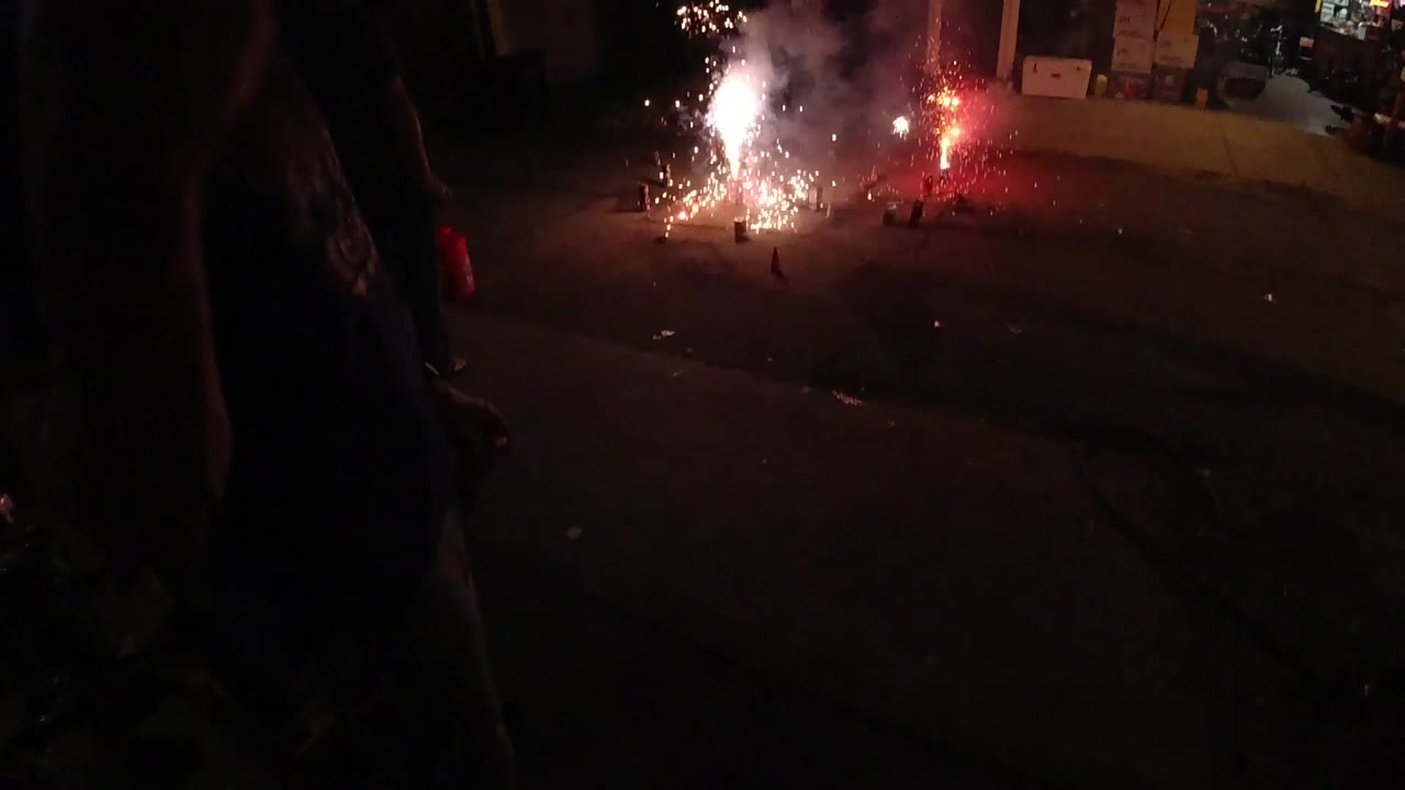 A red flare glows beside a white sparkling firework on the pavement, with smoke drifting upward and an open garage visible across the street or driveway.