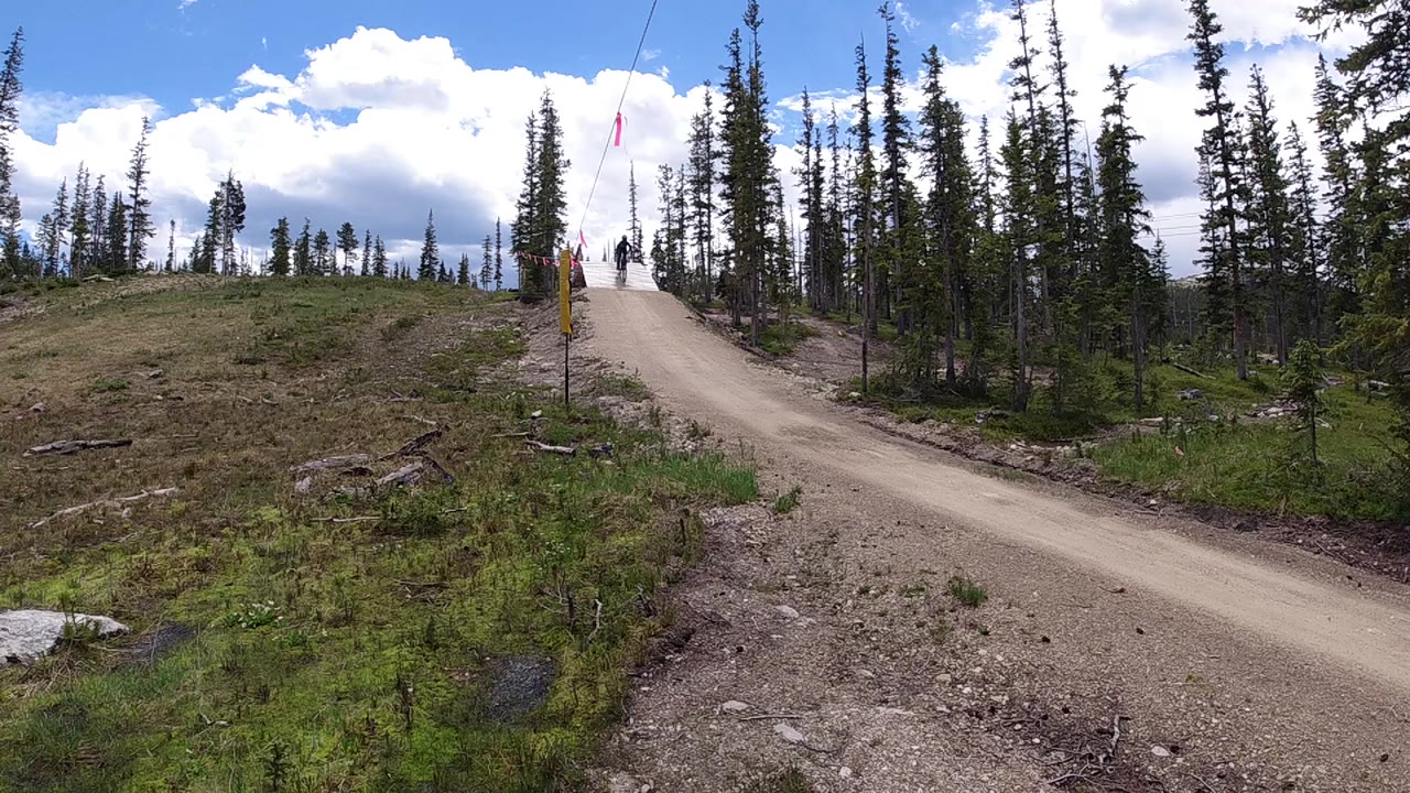 Aerial drone view of a winding dirt single-track trail climbing through sparse alpine conifers, with open rocky terrain on either side and blue sky with white clouds; a possible trail sign or post is visible near the top of the rise; no people visible.
