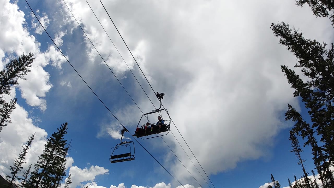 The occupied chairlift sits high on the diagonal cable path, with an empty chair behind it, white cloud cover filling the right side, and treetops framing the lower edges.