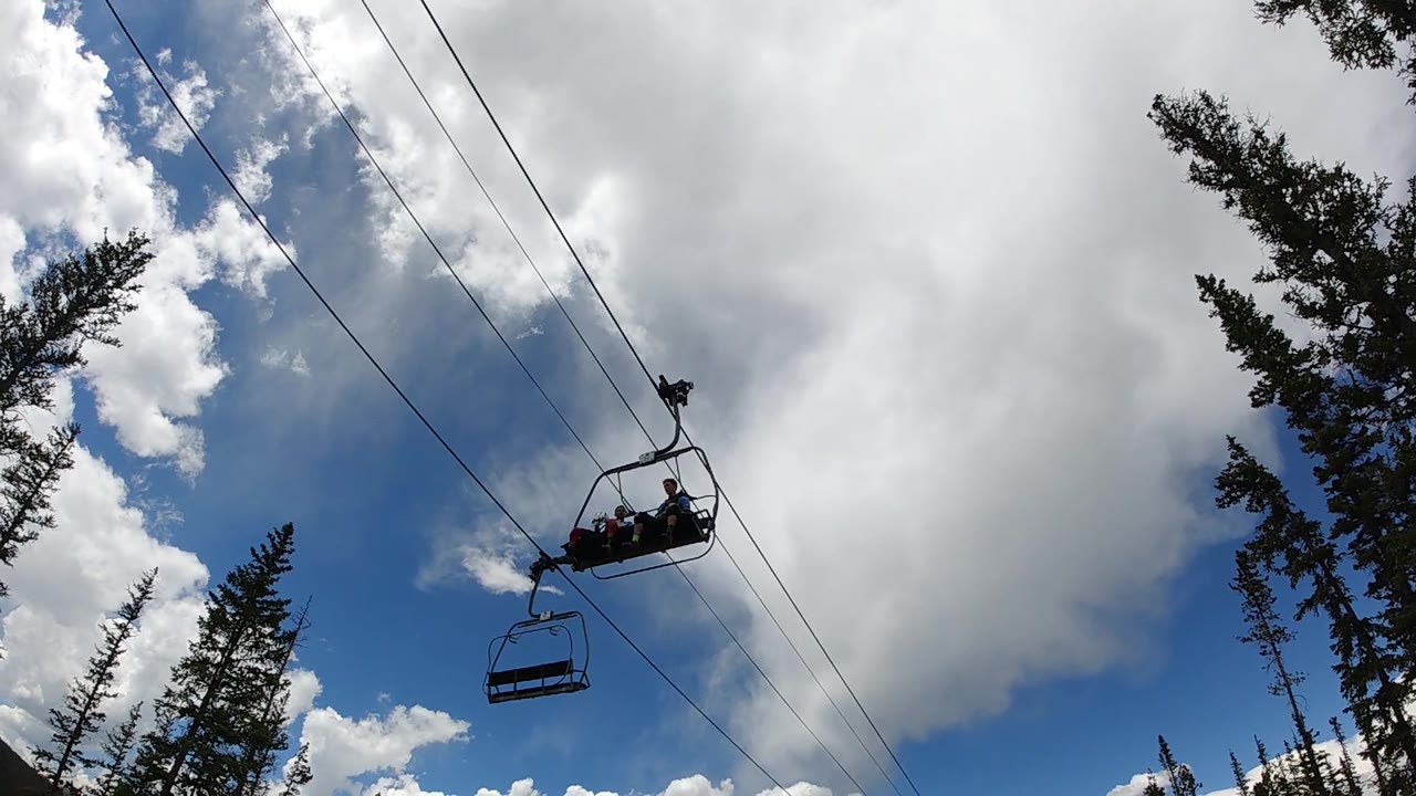 Riders on an open chairlift appear as dark figures near the upper right, with diagonal cables, a trailing empty seat, and tall evergreens bordering the sky.