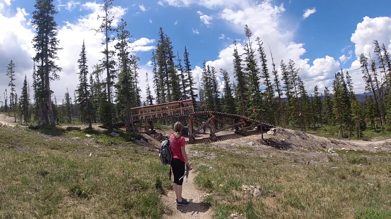 At the opening moment, a woman in a red top and backpack walks away on a narrow dirt path toward a wooden bridge, with tall conifers and bright clouds beyond.