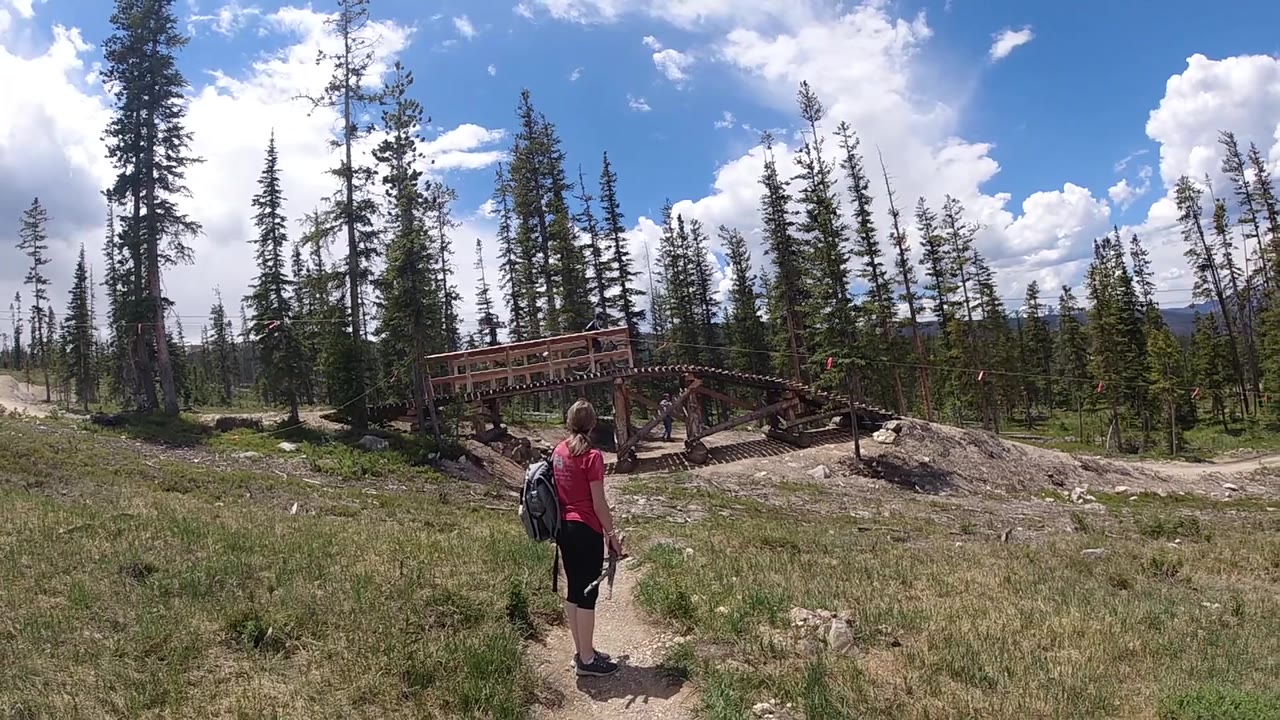 One second in, the backpacker remains near center frame on the narrow trail, with the wooden bridge ahead to the right and a forested ridge under scattered clouds.