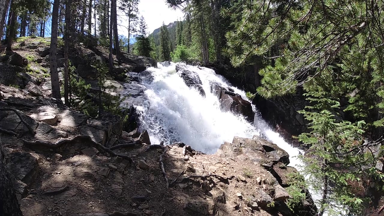 The falls surge across the midground with a narrow stream visible at lower right, shaded rocks in front, and no visible people.