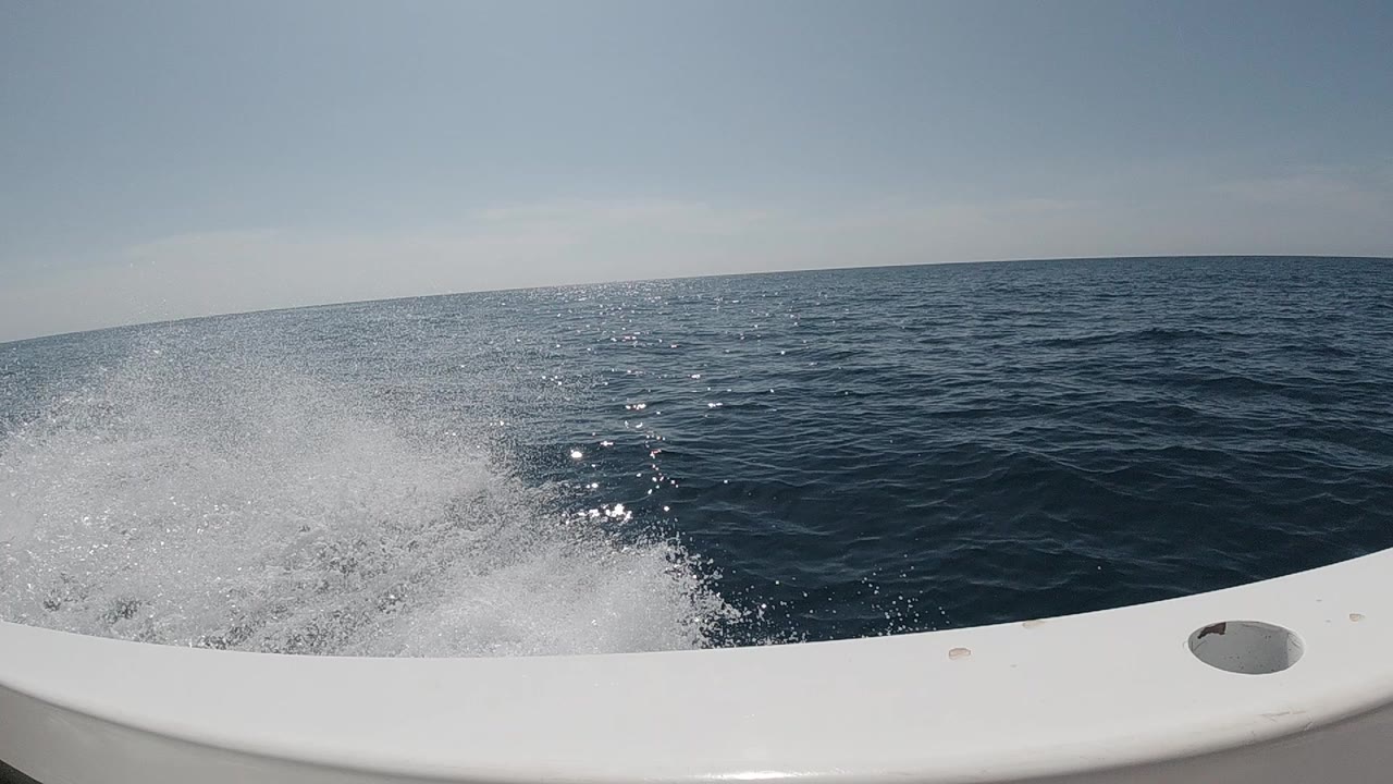 White boat rail crosses the foreground as foamy wake trails over dark blue open ocean, with no visible people in this frame.