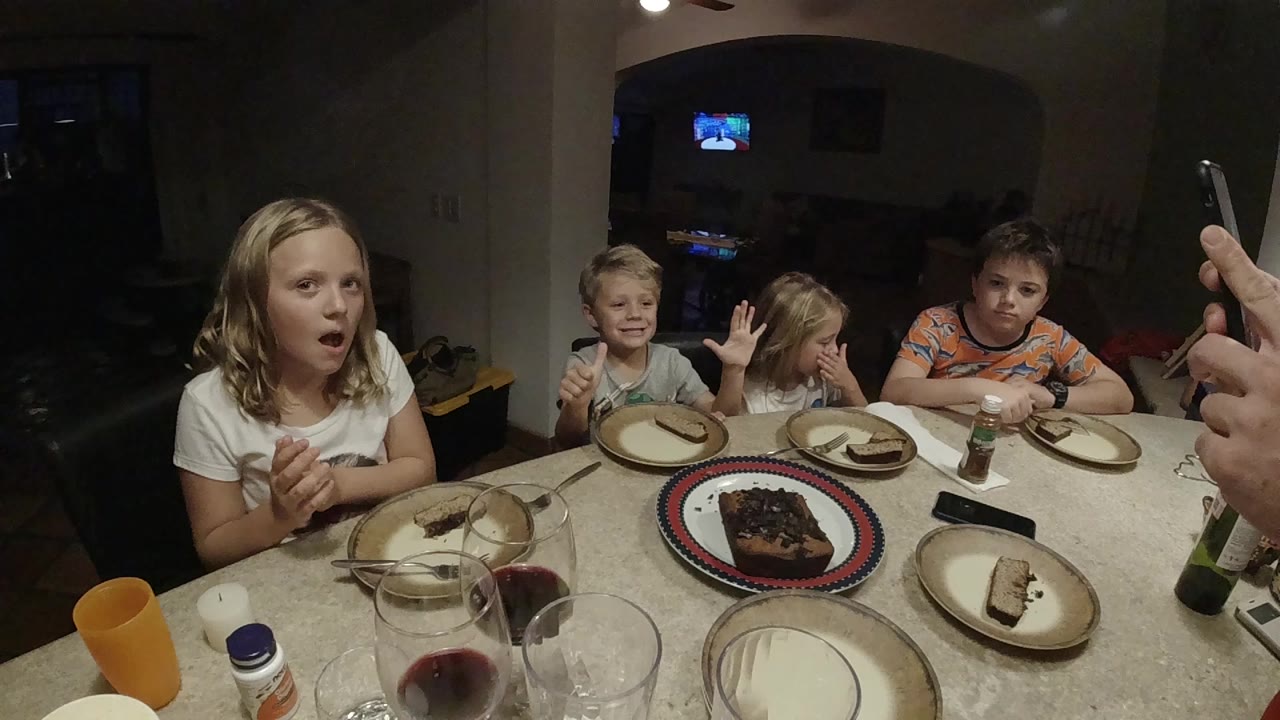 Four children sit around a dining table with a cake plate centered on the table and an adult hand holding a phone at the right edge.
