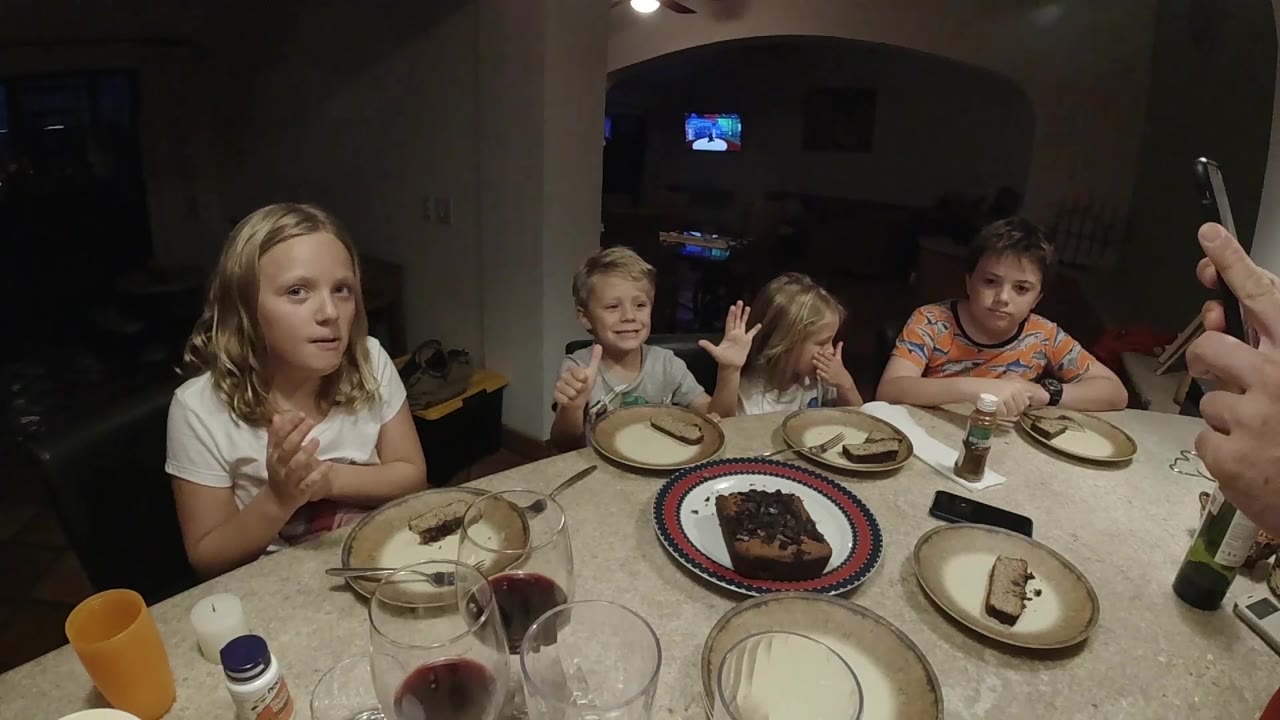 The children look toward the table and camera area, with the central cake plate, empty plates, glasses, and a small dark object on the tabletop.