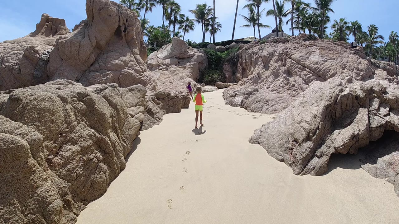 Drone-angle aerial view looking down over large smooth tan rock formations and white sand on a tropical beach; a lone person in yellow-orange clothing stands in the center of the frame with dense palm trees and blue sky visible behind them. No other people are visible.