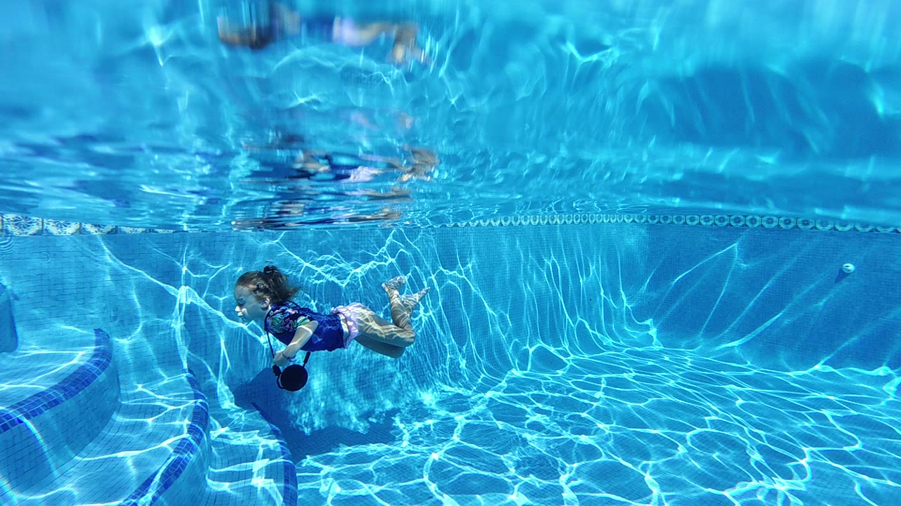 A child in a blue shirt swims underwater near the left side of a clear pool, with sun ripples across the water and the surface line near the top.