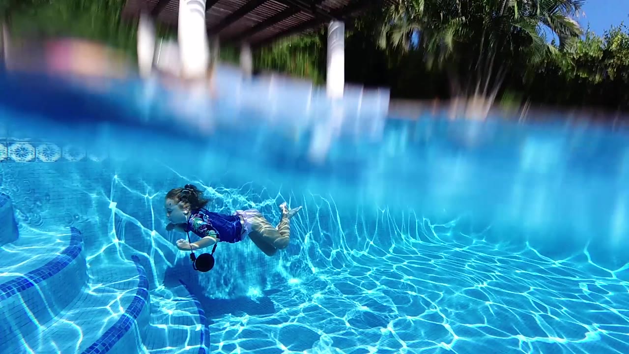 A split-level view shows the child underwater at mid-right, with the rippled surface cutting across the frame and patio greenery blurred above.