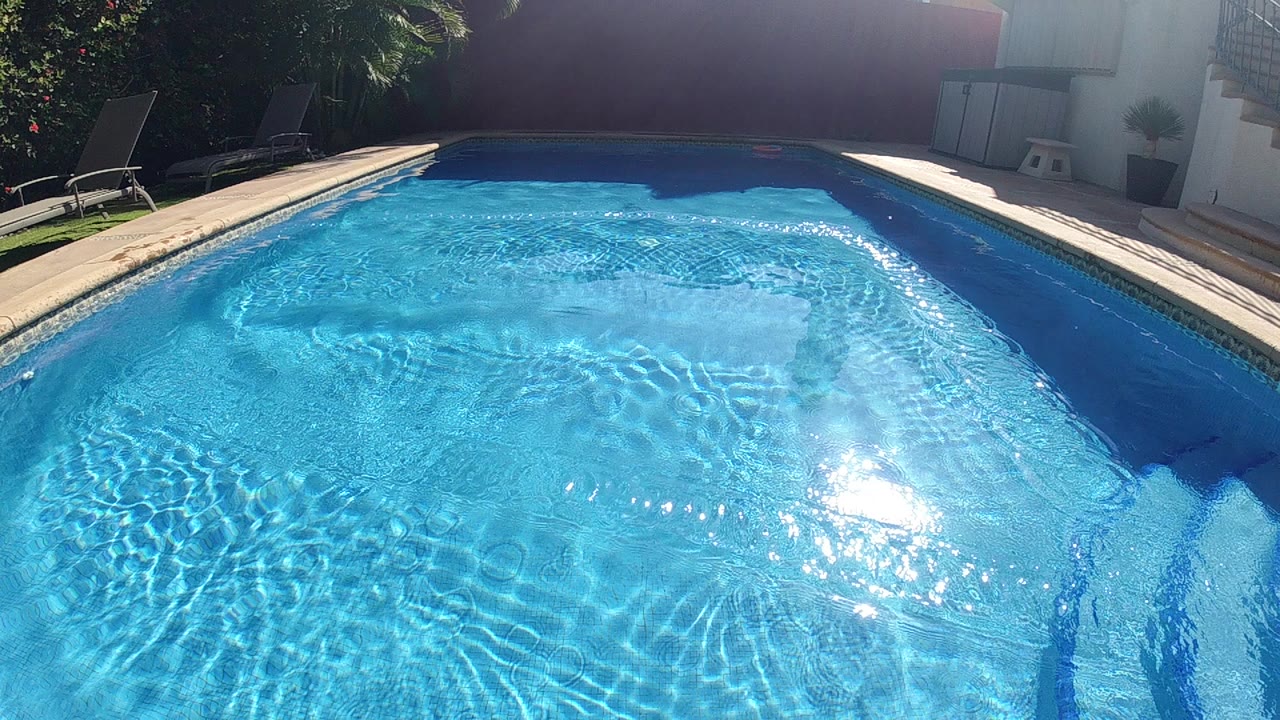 High overhead view of an outdoor swimming pool with vivid blue water filling most of the frame, pale tan pool coping along the lower portion, pool entry steps at lower left, sun reflection glare near upper-left water surface, faint foliage at top corners, no visible people