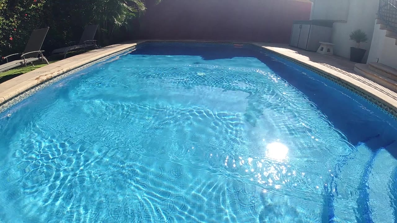 High overhead view of outdoor swimming pool, vivid blue water with bright sun glare near center of the surface, pale tan pool coping across the lower frame, pool steps at lower left, no visible people