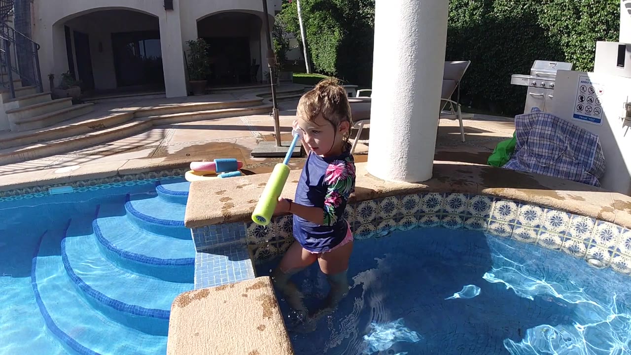 At the start, a child stands chest-deep near the pool edge holding a green water toy beside blue steps and a white column.