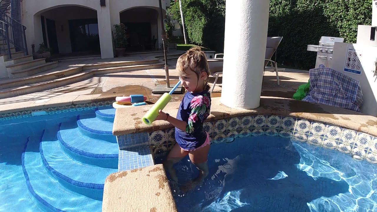The child turns slightly toward the pool while aiming the green toy over the water, with sunlit blue steps to the left.