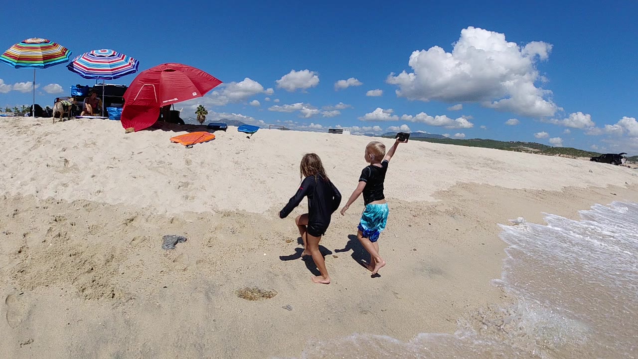Two children walk away from the camera on a Los Cabos beach, with a red umbrella, striped umbrellas, pale sand, and a narrow band of surf at the right edge.