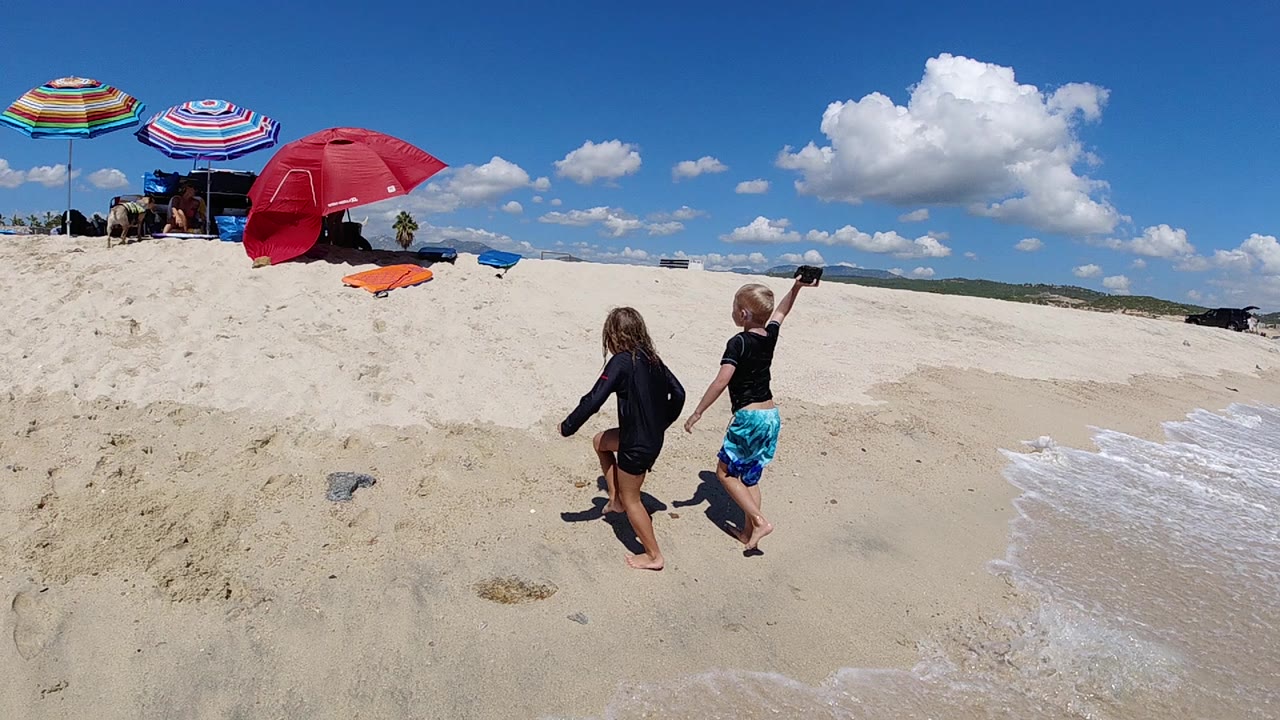 The children continue across the sand in Los Cabos, with one child raising an arm while the red umbrella and striped shade umbrellas sit ahead on the left.