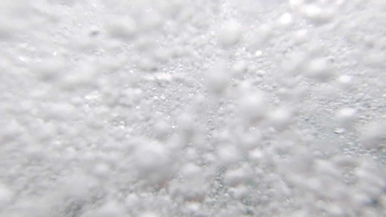 Close-up of white ocean seafoam at the water surface in Los Cabos, Mexico, dense bubble clusters with teal-gray water showing in gaps; no people visible
