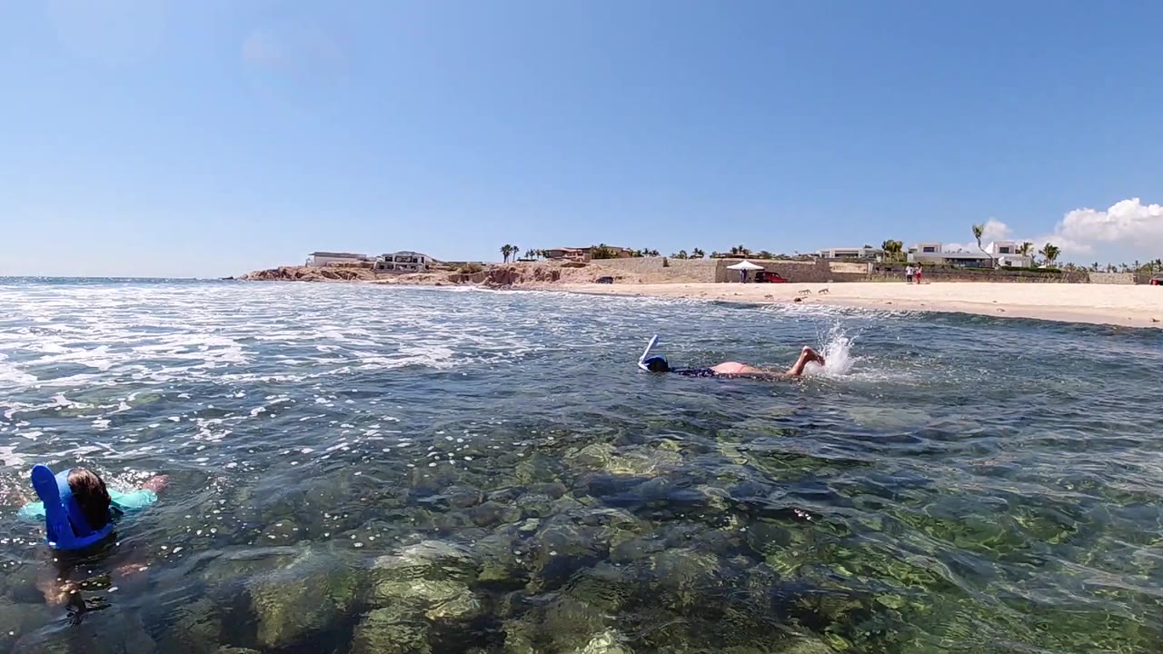 Two snorkelers move through clear shallow water in Los Cabos, with rocks visible below, white surf at left, and sandy beach behind.