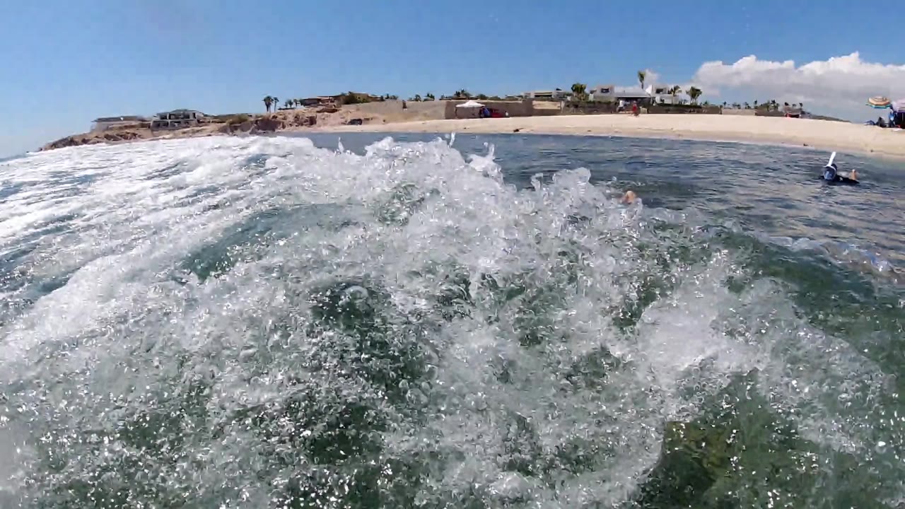White foam breaks across the water-level view in Los Cabos, Mexico, while the sandy shoreline and low coastal structures remain visible in the distance.