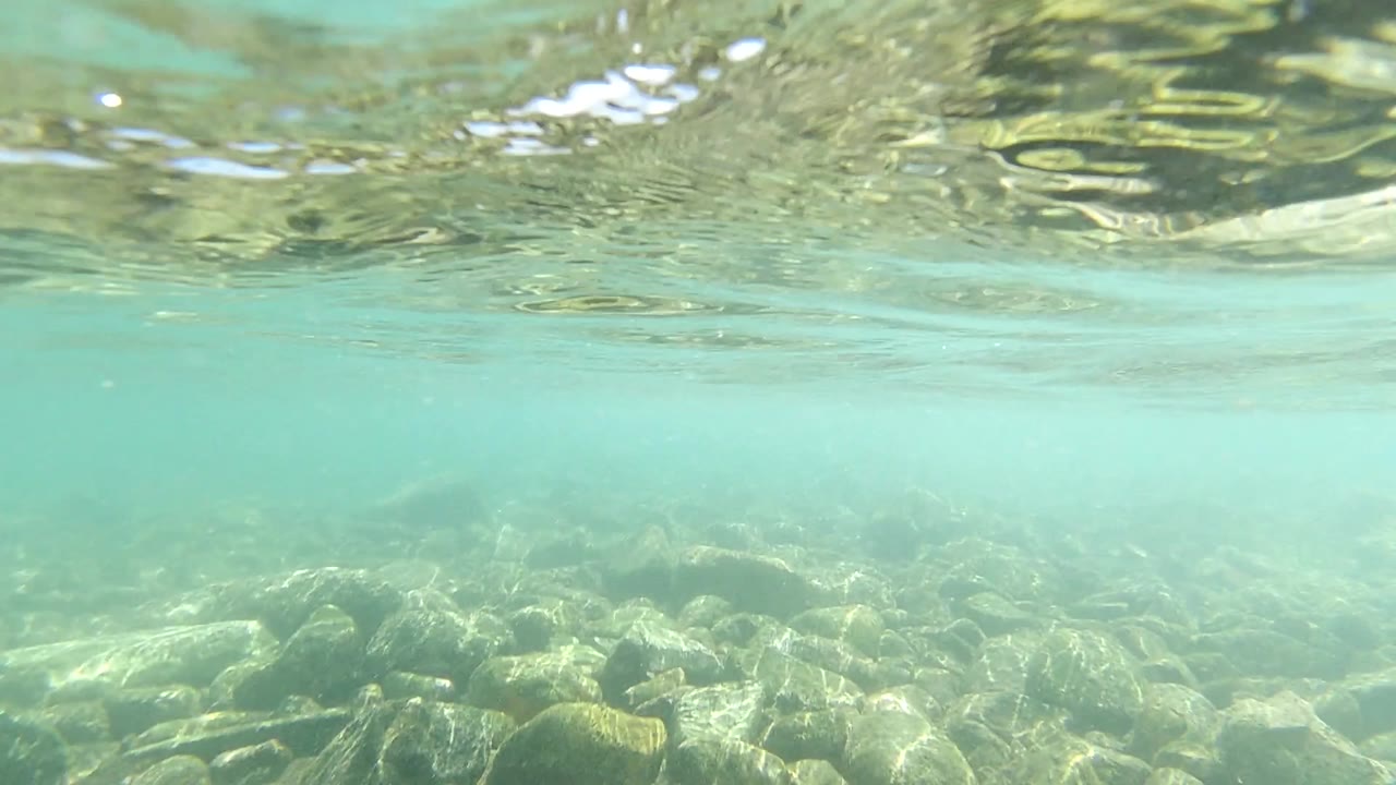 Early underwater frame in Los Cabos showing pale turquoise water, a rippled surface band, rounded rocks on the seabed, and no visible people.