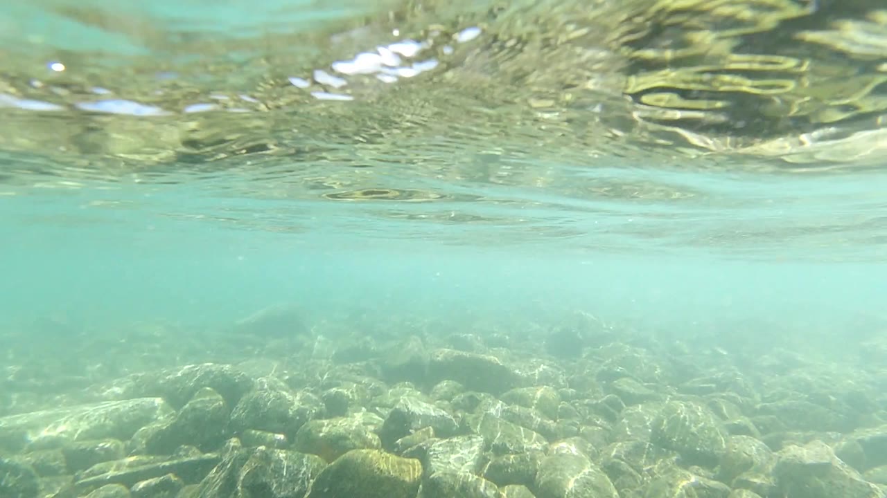 Los Cabos underwater frame with the surface shimmering across the top, soft haze in the middle water, rounded rocks below, and no visible people.
