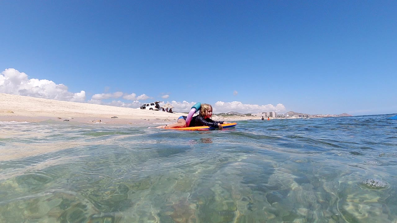 A child on a bright inflatable float is centered in shallow turquoise water near a sandy Los Cabos shoreline under blue sky.