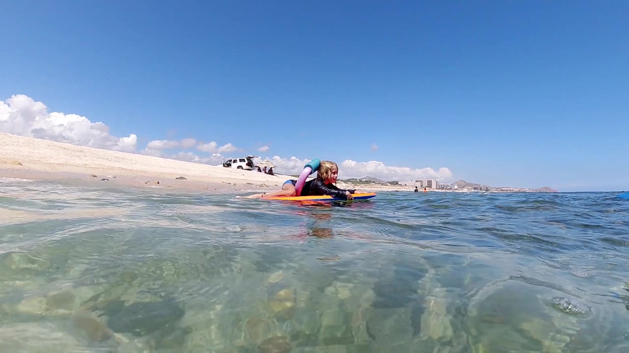Small waves rise across the foreground while the child on the inflatable faces the camera near the Los Cabos beach.