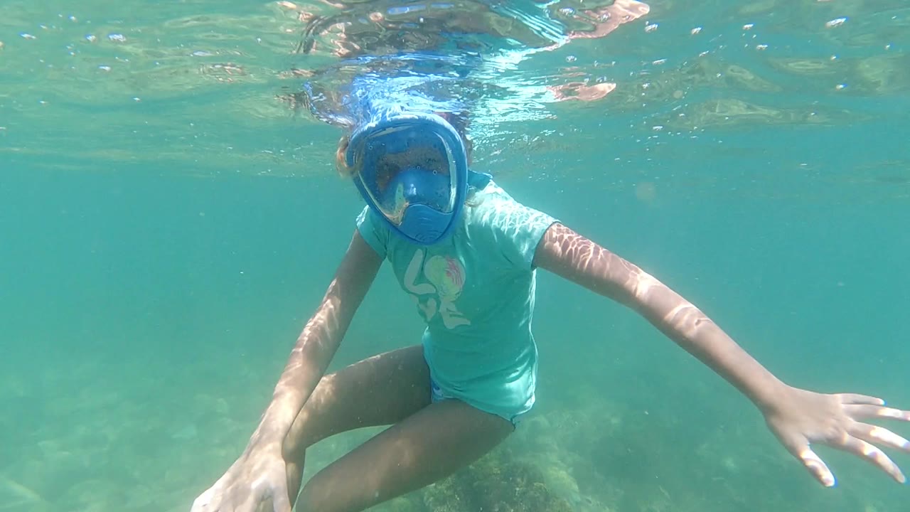 A woman in Los Cabos floats just below the water surface with a blue snorkel mask, turquoise shirt, and one arm reaching across the clear water.