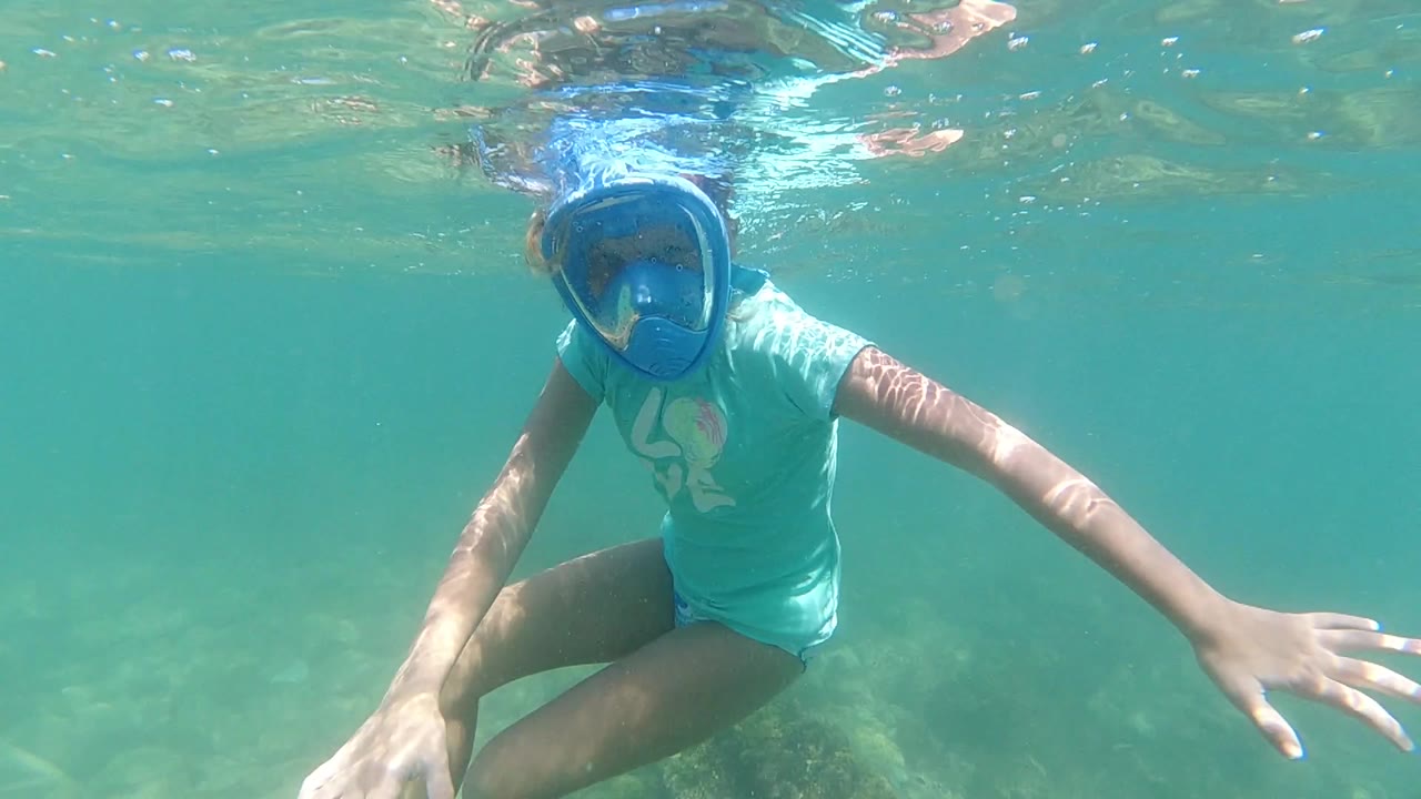 The Los Cabos snorkeler faces forward under rippled surface water, with her right arm extended and pale rocks visible behind her legs.