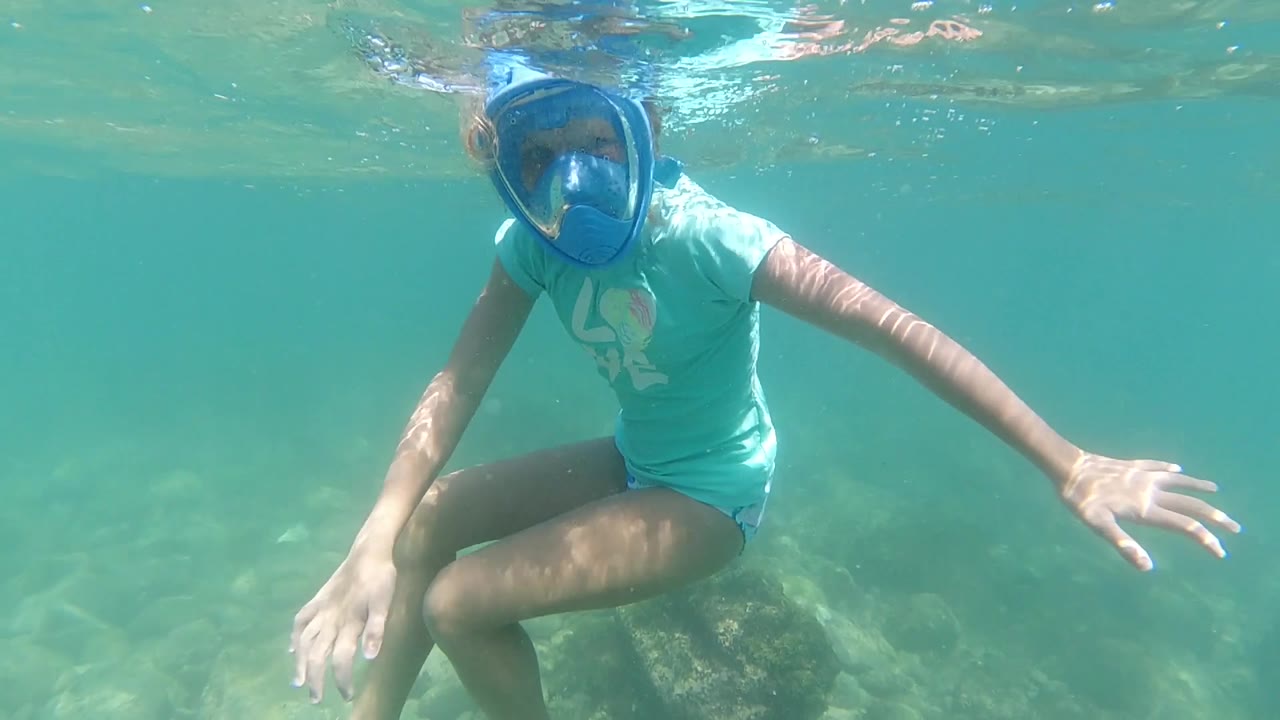 A woman in a blue mask hovers underwater in Los Cabos with one knee bent, one hand open, and sunlight wavering across the shallow rocks.