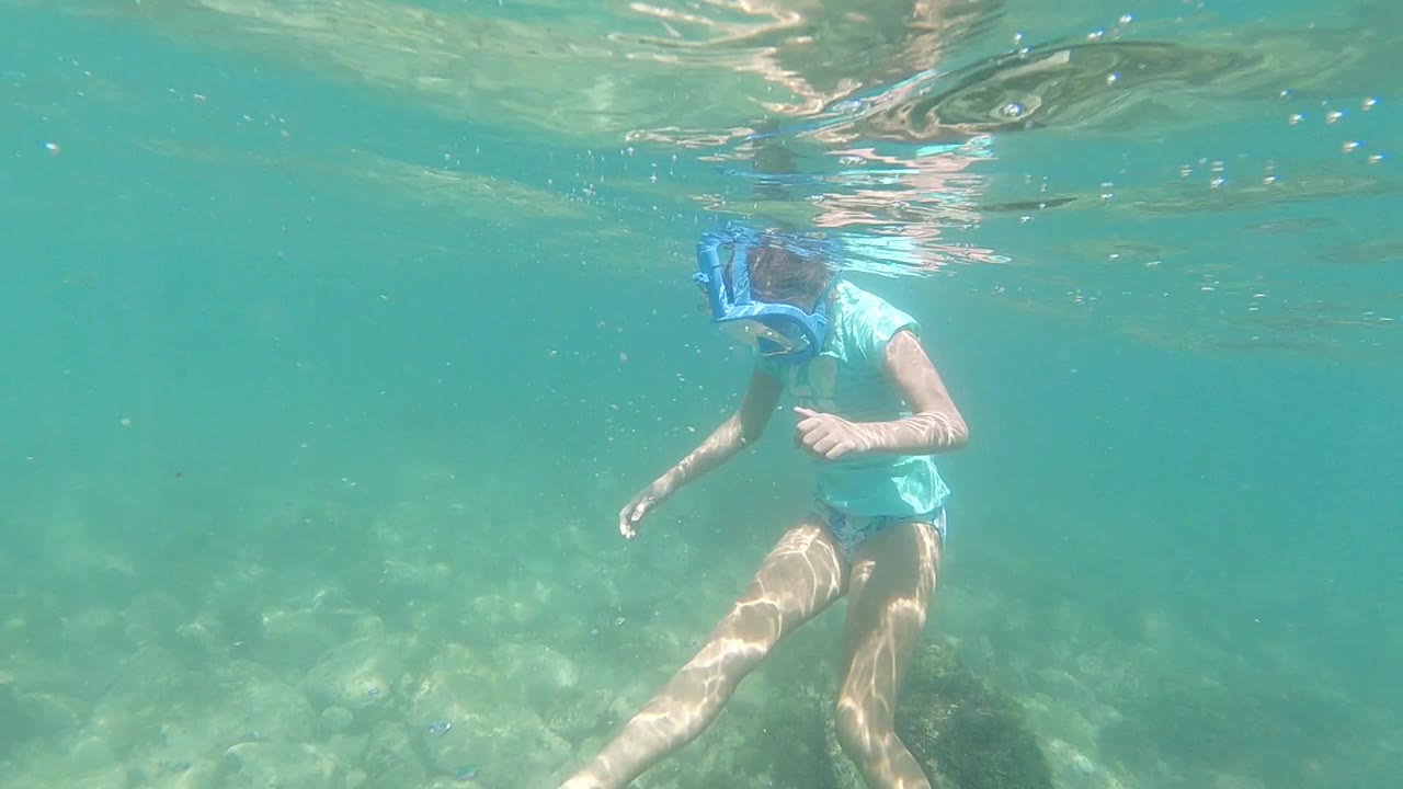 The woman remains close to the surface with her snorkel mask at the top of frame and one leg extended over the sunlit rocks below.