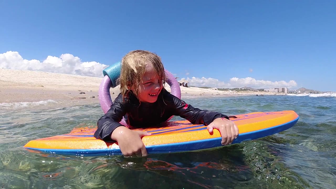At 0:00, a young girl grips an orange and blue bodyboard at water level in Los Cabos, Mexico, with sandy beach and white clouds behind her.
