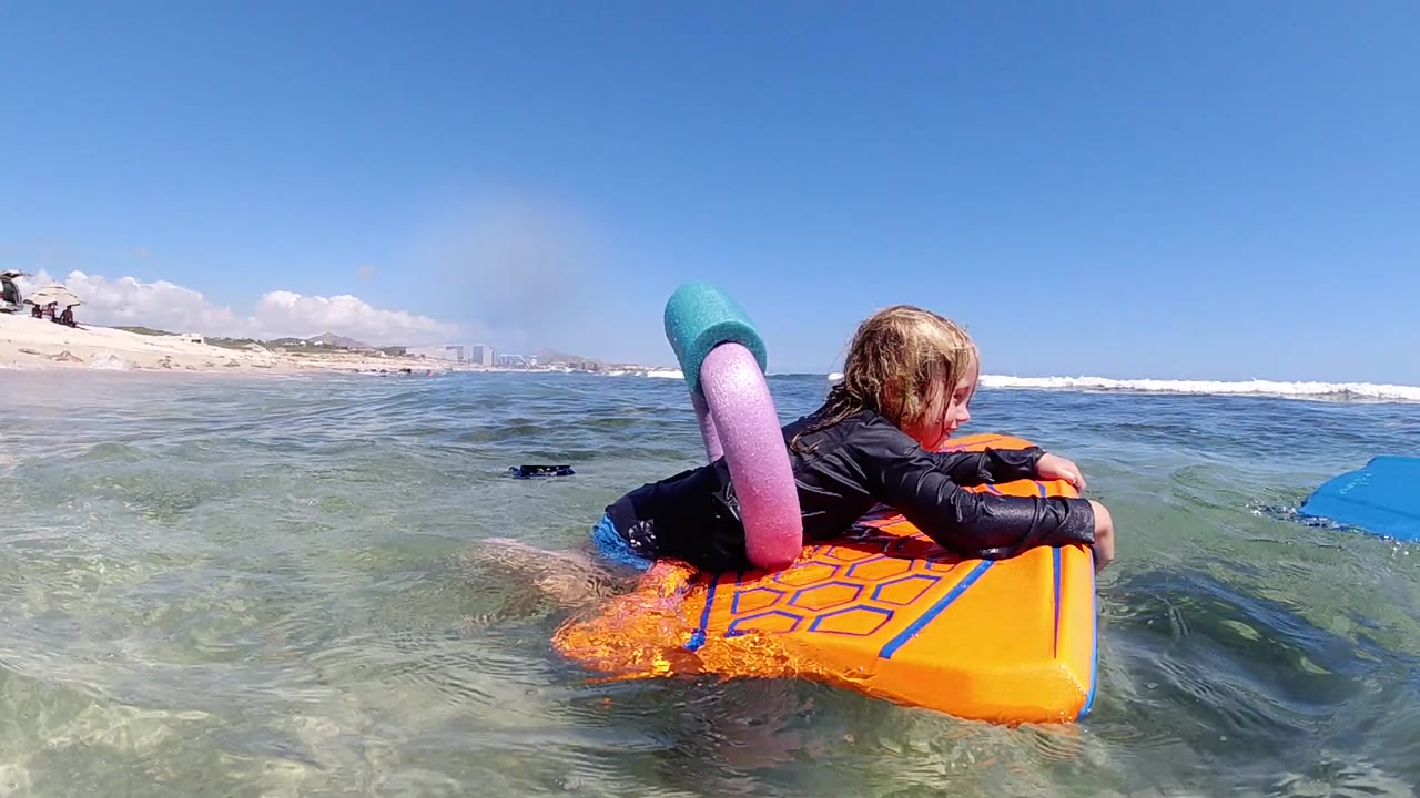 At the start of the Los Cabos clip, a blonde young girl rests on a bright orange float in clear shallow water with surf and sandy shoreline behind her.