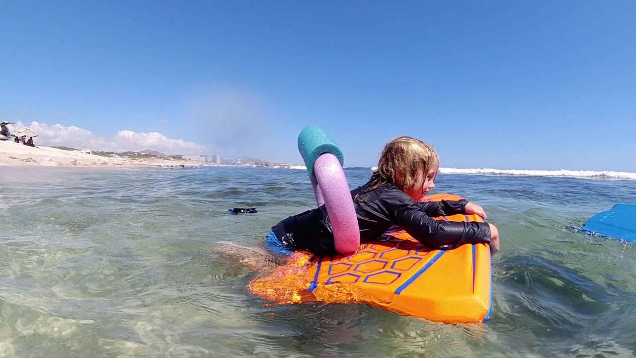 One second in, the girl faces slightly away while holding the orange float, with rippled turquoise water and the low Los Cabos shoreline in the background.