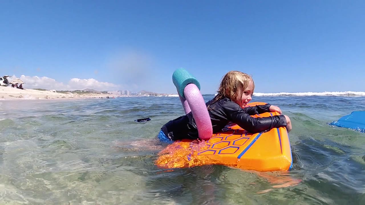 The girl turns her face toward camera while lying on the orange float, with a small swell passing through the shallow Los Cabos water.