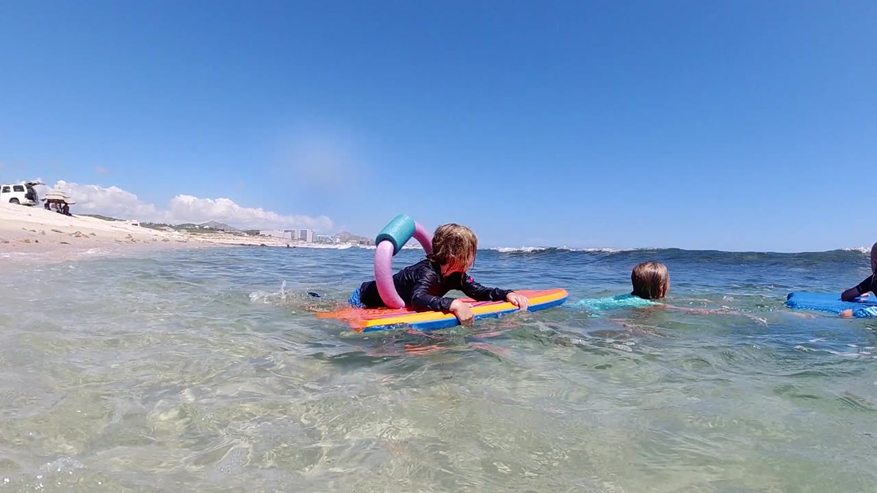 At the start of the Los Cabos, Mexico clip, a child lies on a bright board in clear shallow water while another child swims nearby and the sandy beach sits at left.