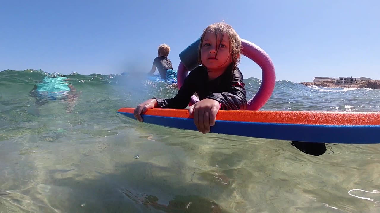 The girl stays centered on the flotation device as a small wave rises beside her and another person floats farther back in the Los Cabos water.