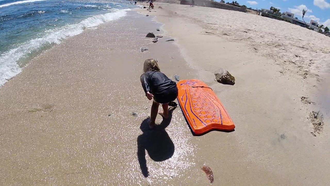 Little girl bends beside an orange floater on a sandy beach.