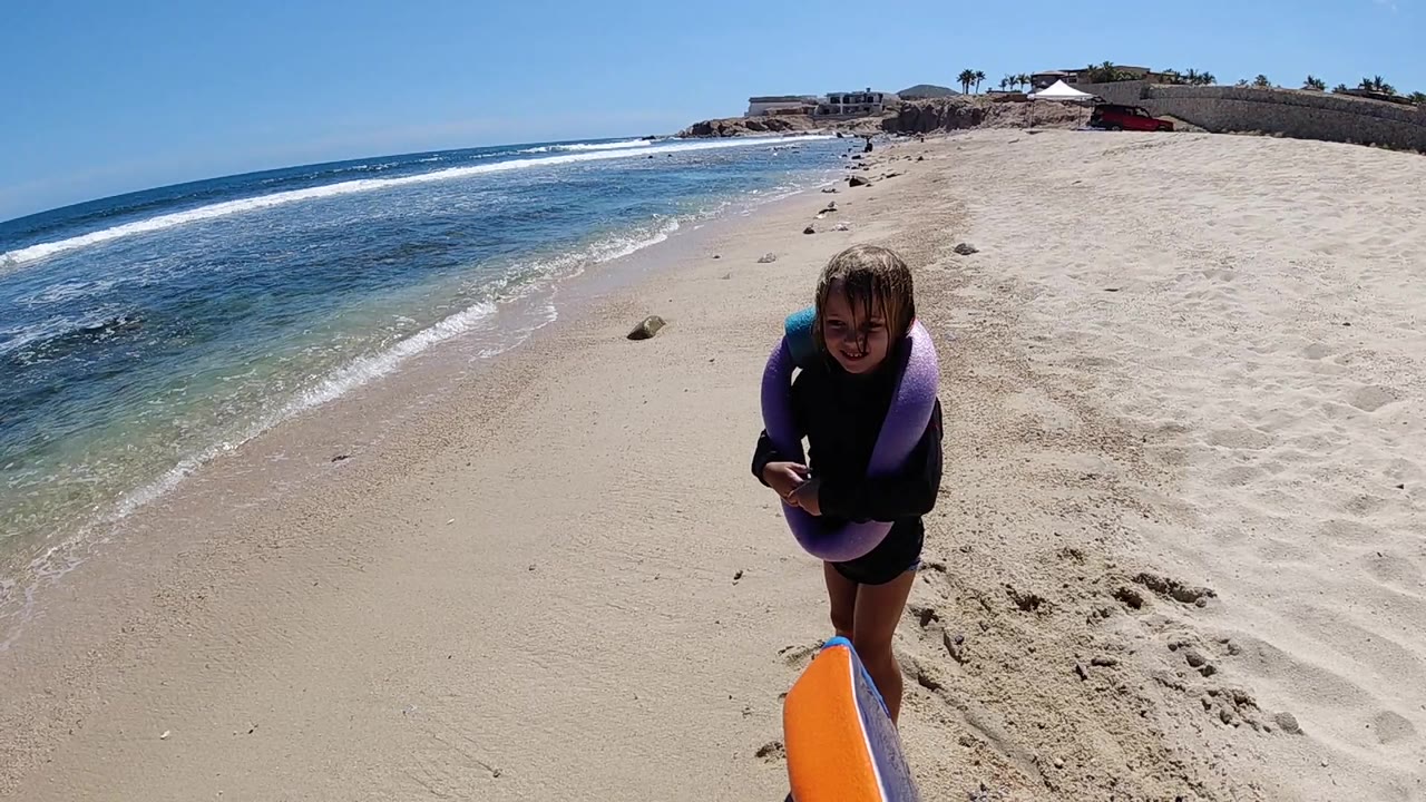 A young girl in a purple flotation aid stands near an orange board on a Los Cabos beach, with blue ocean and white surf stretching along the left side.