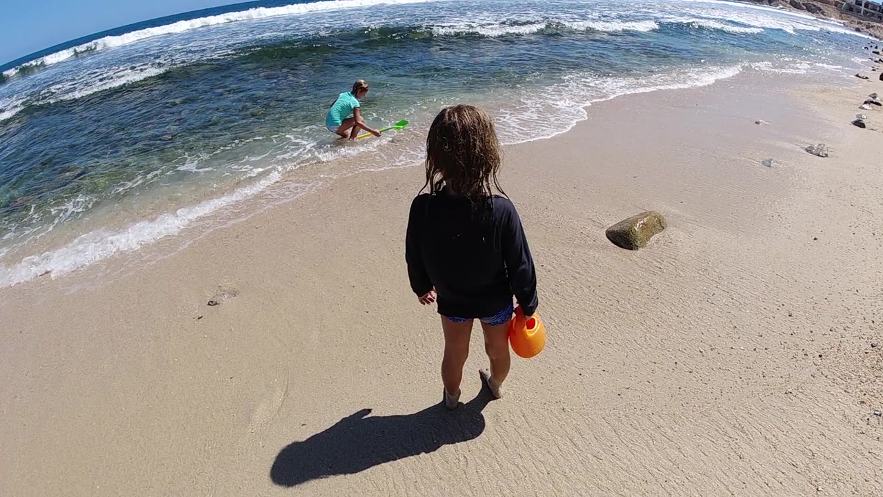 At the start of the Los Cabos clip, a young girl stands on bright sand with an orange bucket while an adult in turquoise crouches near the foamy surf.