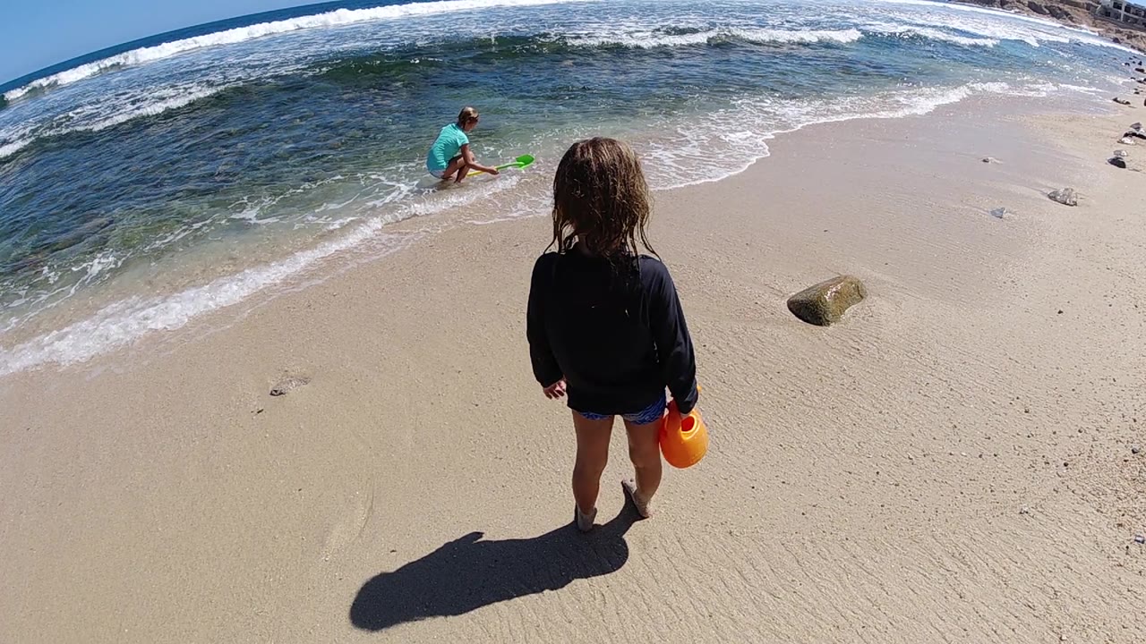 Child watches from dry sand while small waves wash behind an adult on the beach.