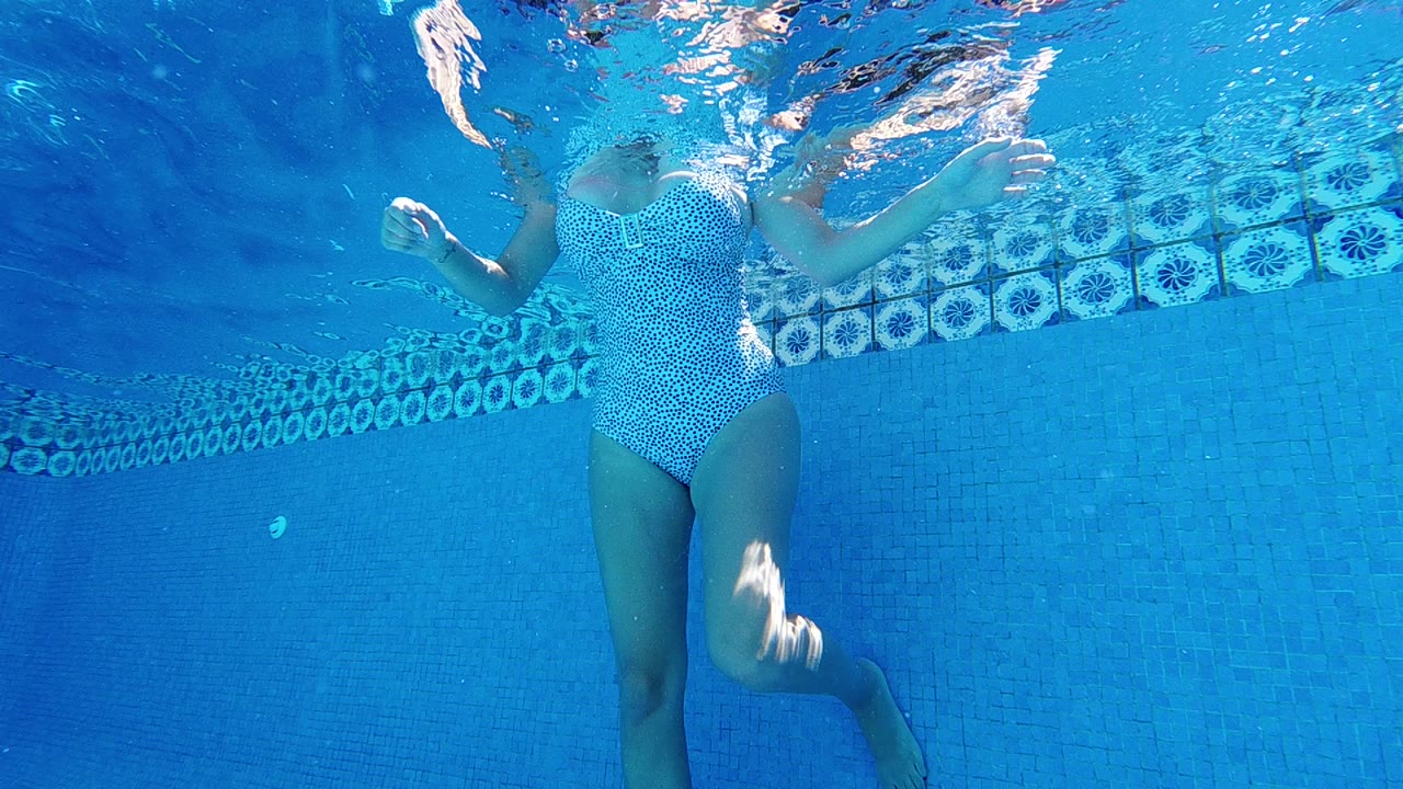 A woman in a white spotted swimsuit is underwater near a blue swimming pool wall, with her bent leg and arms visible below the rippled surface.